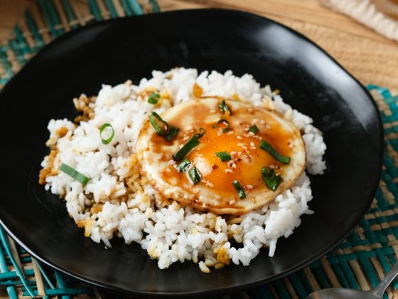 Easy egg recipes. An image of a Korean style egg rice bowl. there is rice inside a black bowl on top of a table with a sunny side up egg and soy sauce on top with sprinkled spring onions.