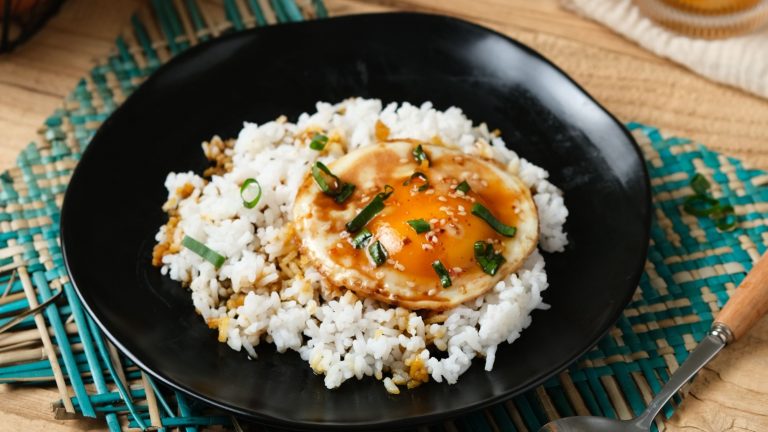 Easy egg recipes. An image of a Korean style egg rice bowl. there is rice inside a black bowl on top of a table with a sunny side up egg and soy sauce on top with sprinkled spring onions.